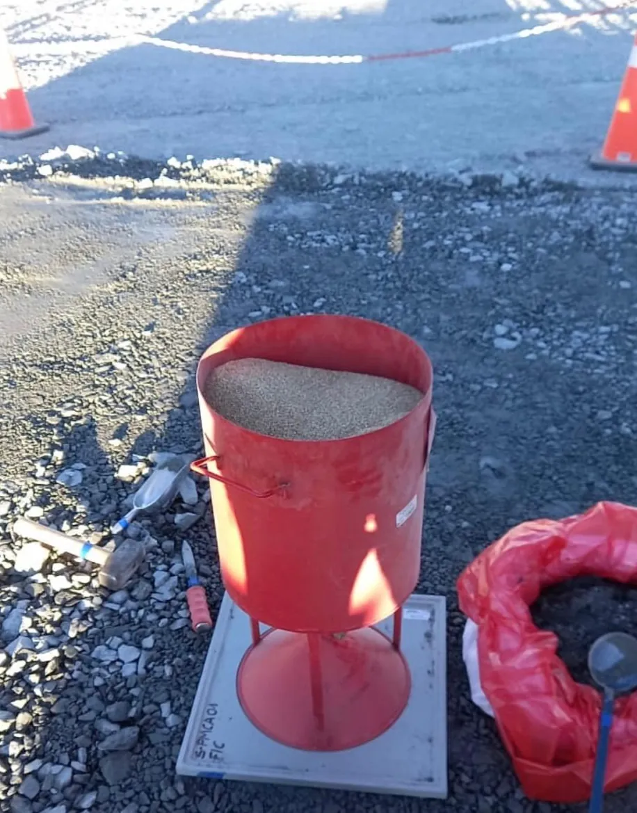 Equipo de cono de arena cargado con arena sobre su placa base, junto a una bolsa de muestras